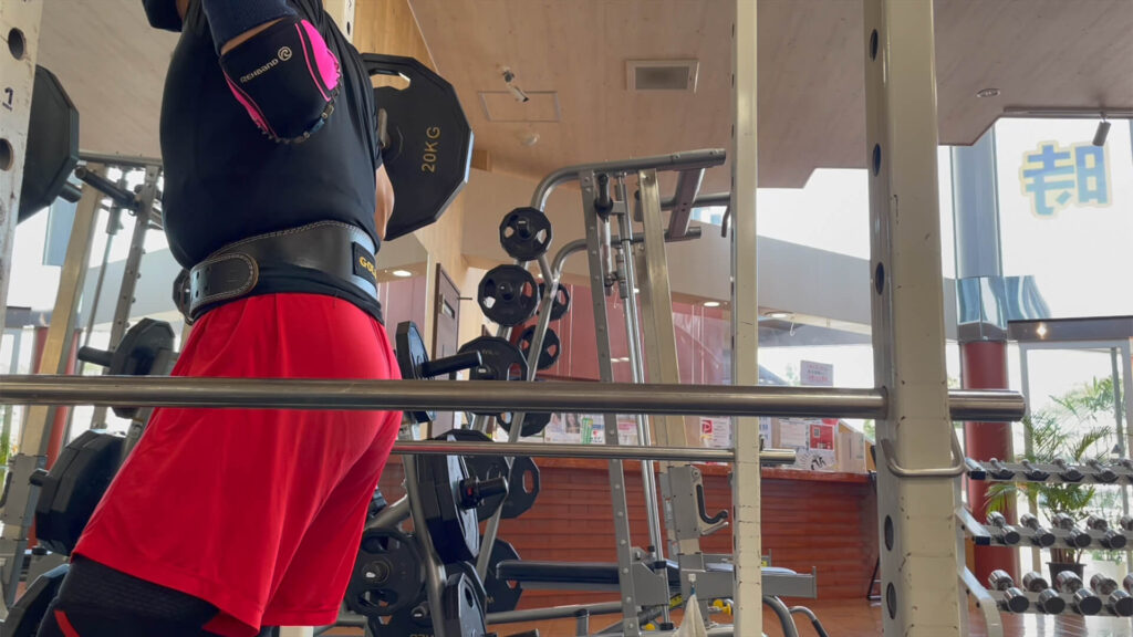 長崎県南島原市で店舗数最多のダイエット教室・健康体操クラブであるZUTTOWAKAIの代表である太田祥平がダイエットやメタボ改善に効果的なスクワットやり方を実演する様子。膝を軽く曲げてバーベルの真下にカラダを入れている