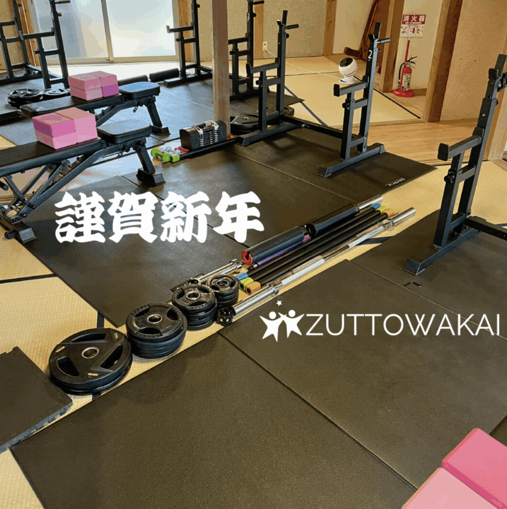 フィットネスクラブZUTTOWAKAIのトレーニング施設内に設置された筋力トレーニングエリアの様子。畳敷きの室内に黒いトレーニングマットが敷かれ、ベンチ台、スクワットラック、バーベル、プレート、ダンベルなどの器具が整然と並ぶ。ピンク色のクッションパッドも目立つ位置に配置。左側には白抜き文字で「謹賀新年」、右下にはZUTTOWAKAIのロゴが大きく表示されており、新年の挨拶ビジュアルとして作成されたことがわかる。