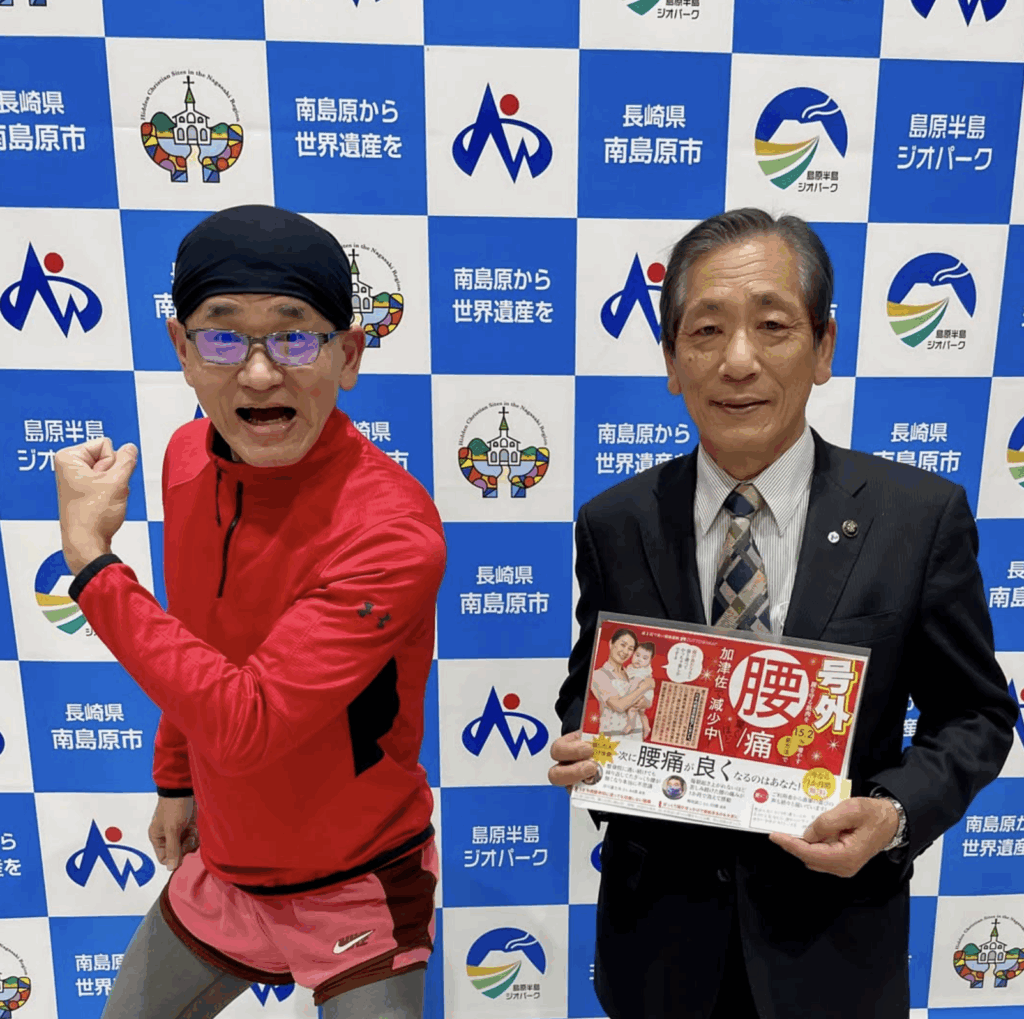 ZUTTOWAKAI代表の男性(赤いスポーツウェア姿)と南島原市の松本政博市長(スーツ姿)が並んで写っている記念写真。背景には「長崎県南島原市」「南島原から世界遺産を」「島原半島ジオパーク」などのロゴが並んだパネル。松本市長は「腰痛改善プログラム」の案内チラシを手に持ち、ZUTTOWAKAIの活動をPRする場面。