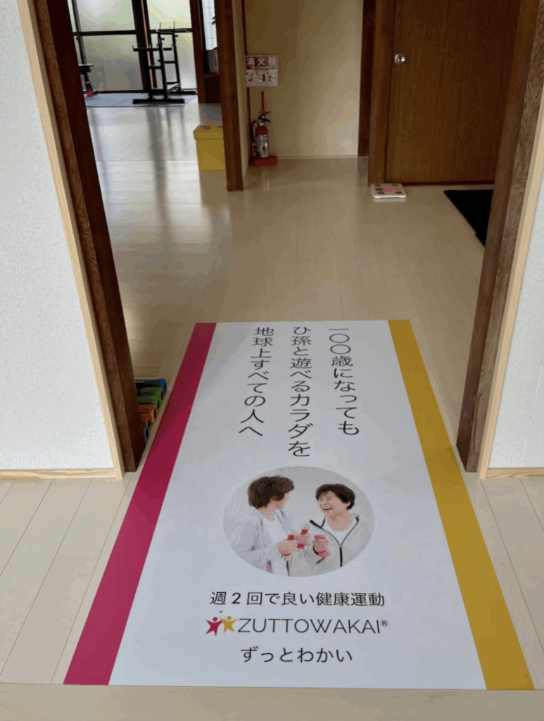 ZUTTOWAKAI南島原南有馬の施設内の様子。明るい木目調のフロアに、「100歳になってもひ孫と遊べるカラダを地球上すべての人へ」と書かれた大型マットが敷かれている。マットの中央には笑顔でプロテインを手にする高齢女性2人の写真と、「週2回で良い健康運動 ZUTTOWAKAI ずっとわかい」のロゴ入りメッセージ。奥の部屋には筋トレ機器や消火器、カラフルなストレッチポールなどが配置されている。
