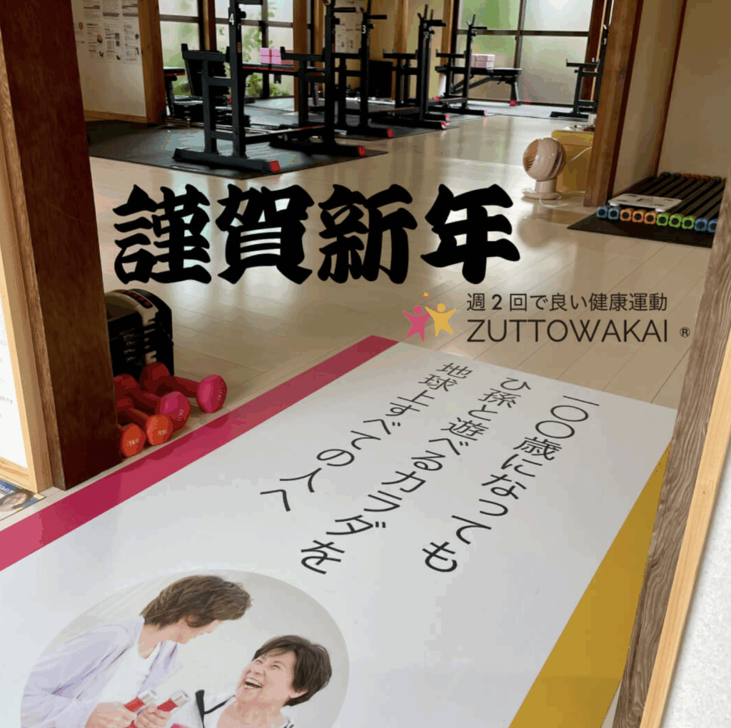 ZUTTOWAKAIのトレーニング施設内に飾られた2024年の新年あいさつビジュアル。床には「100歳になってもひ孫と遊べるカラダを地球上すべての人へ」というスローガンと、笑顔の高齢女性2人の写真が印刷された大型マットが敷かれている。奥には筋力トレーニング用のラックやダンベルが並び、中央には「謹賀新年」と大きく黒文字で表示され、右下にはZUTTOWAKAIのロゴと「週2回で良い健康運動」の文字が添えられている。