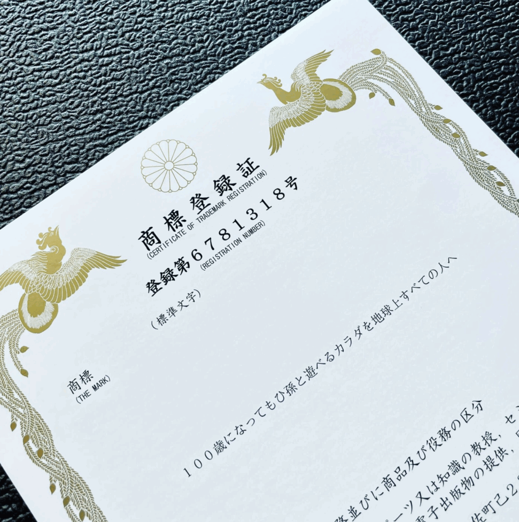 日本の特許庁が発行した商標登録証の写真。上部には金色の鳳凰と菊の紋章がデザインされ、中央には「商標登録証」「登録第6781318号」と記載。商標(標準文字)は「100歳になってもひ孫と遊べるカラダを地球上すべての人へ」。白地に黒文字で印字され、高級感のある装丁が特徴。