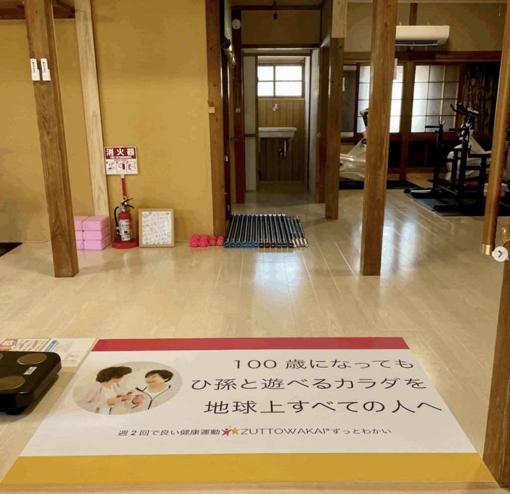 ZUTTOWAKAI南島原口之津の玄関から見た室内の様子。正面には木造の柱と和の内装が特徴の空間が広がり、奥には運動器具(エアロバイクなど)や洗面所が見える。床にはカラフルなバーベルやダンベルが並び、左手前には体組成計が置かれている。床の中央には「100歳になってもひ孫と遊べるカラダを地球上すべての人へ 週2回で良い健康運動 ZUTTOWAKAI」と書かれたメッセージマットが敷かれている。