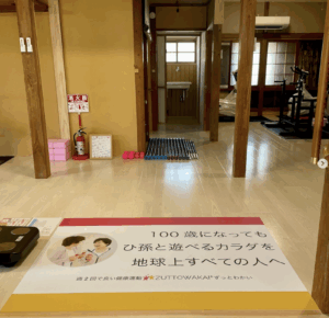 ZUTTOWAKAI南島原口之津の玄関から見た室内の様子。正面には木造の柱と和の内装が特徴の空間が広がり、奥には運動器具（エアロバイクなど）や洗面所が見える。床にはカラフルなバーベルやダンベルが並び、左手前には体組成計が置かれている。床の中央には「100歳になってもひ孫と遊べるカラダを地球上すべての人へ　週2回で良い健康運動 ZUTTOWAKAI」と書かれたメッセージマットが敷かれている。