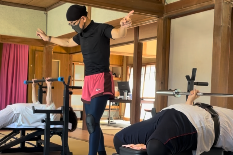 和室のような木造の空間で、ベンチプレスのトレーニングを行うシニア世代の人々。中央にはインストラクターが立ち、正しいフォームを指導している。