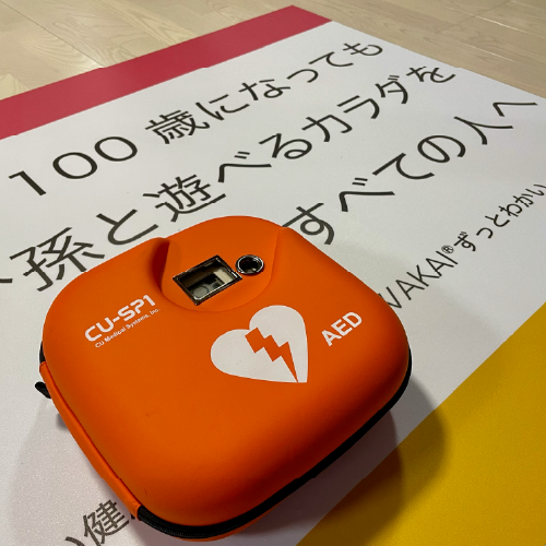 「100歳になっても孫と遊べるカラダを すべての人へ」と書かれたZUTTOWAKAIのスローガンポスターの上に、オレンジ色のAED（自動体外式除細動器）が置かれている写真。AEDは「CU-SP1」モデルで、白い心臓と電撃マークが描かれたコンパクトなケースに収納されている。安全への配慮と、シニア世代の健康支援を掲げるクラブの理念が伝わる構図。
