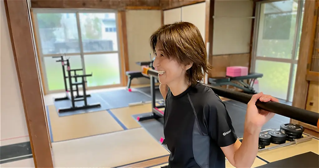 和室風のトレーニングジムで、黒いウェアを着た中年女性が笑顔でスクワットの姿勢をとっている様子。肩にバーを担ぎ、しっかりとしたフォームでトレーニングに取り組んでいる。背景にはトレーニング器具やピンクのヨガブロックが整然と配置されている。