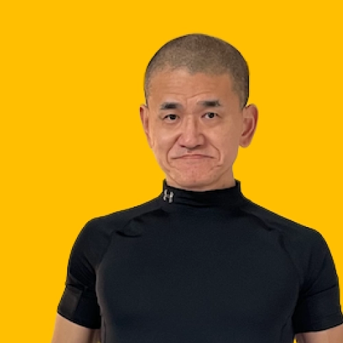 黒のコンプレッションウェア（アンダーアーマーの半袖シャツとタイツ）を着用した中年の男性が、室内でまっすぐ立ってカメラを見つめている。男性は短髪で、軽く笑みを浮かべている。