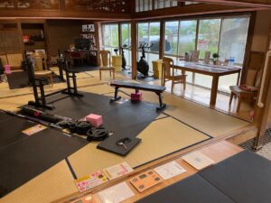 ZUTTOWAKAI 南島原加津佐の筋トレエリア。赤の縁取りが特徴的なトレーニングベンチや、ピンク色のヨガブロックが和室に柔らかく映える。各器具に案内表示や安全のためのピンク色マークが貼られており、丁寧に整備された印象を受ける。
