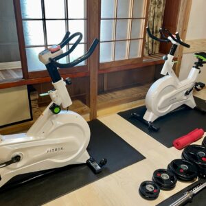 和室の雰囲気を残したトレーニングルームに設置された、FITBOXのスピンバイク2台とダンベル・バーベルのプレート。畳縁のような木製の床と障子窓が、落ち着いた空間に運動のしやすさを調和させている。