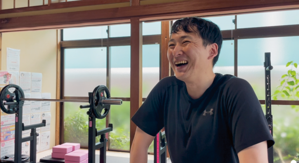 ZUTTOWAKAIの専属運動講師・森川正章が、インタビュー中に笑顔で語る様子。バーベルやトレーニング器具が並ぶ明るい室内で、黒いTシャツ姿の森川さんが楽しそうに笑っている