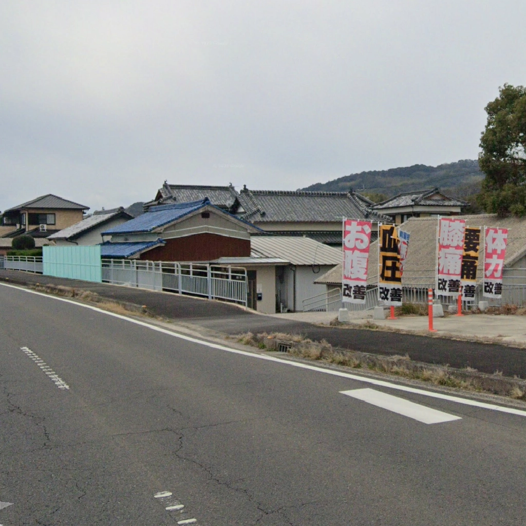 曇り空の下、ZUTTOWAKAI南島原南有馬の入口付近の風景。右手には「血圧改善」「腰痛改善」「体力改善」など、健康に関する赤い文字が書かれたのぼり旗が複数並んでおり、その奥には瓦屋根の住宅地と山並みが広がっている。
