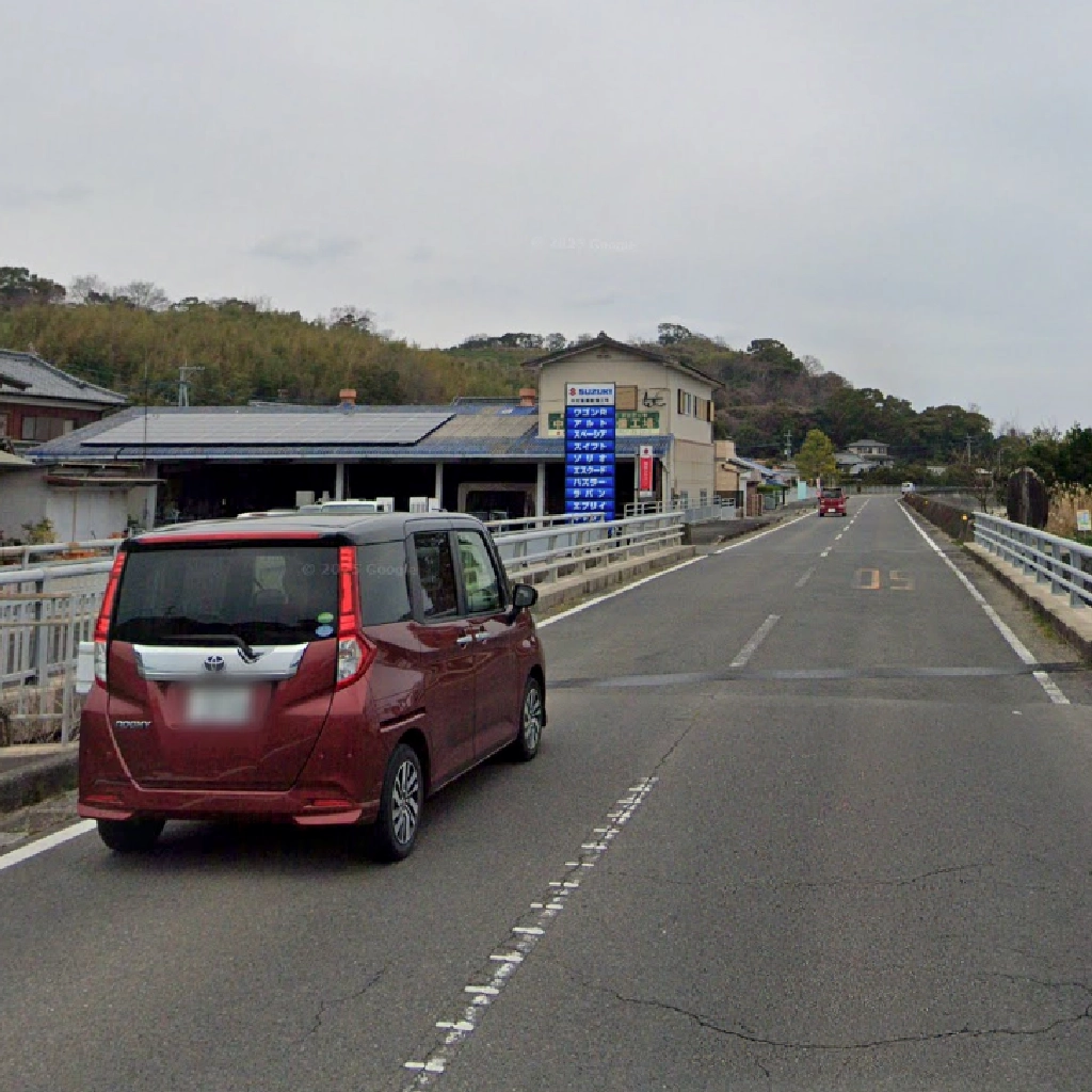 曇り空の下、長崎県南島原市の口之津方面から南有馬方面へ向かう道路の風景。手前には赤色のトヨタ・ルーミーが走行しており、左手には「SUZUKI」の青い看板を掲げた自動車整備工場が見える。