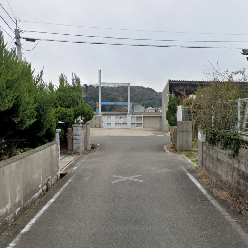 住宅に挟まれた細いアスファルトの路地の先に、口之津港の施設が見える風景。突き当たりには白いゲートのような構造物があり、その背後に山が広がっている。道幅は狭く、両脇はコンクリートの塀や生垣に囲まれている。