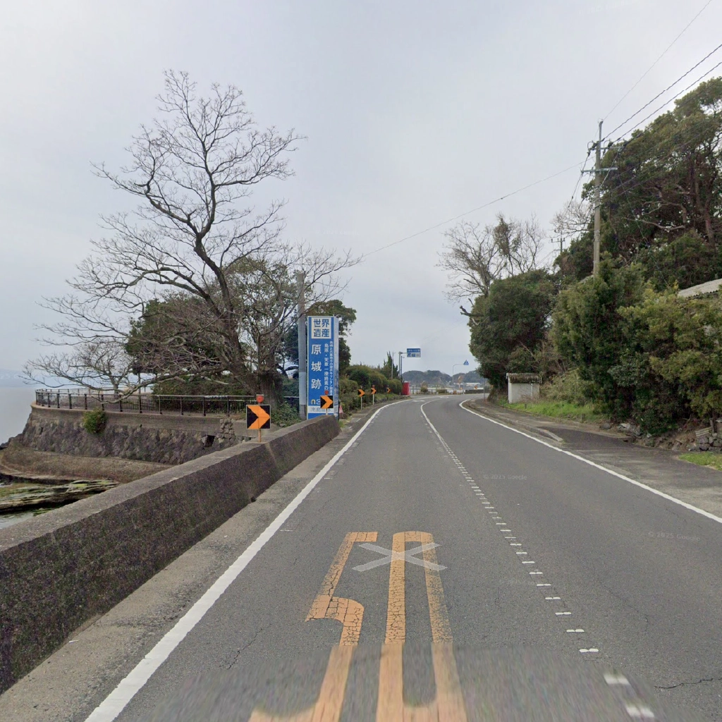 曇り空の下、海岸線に沿って続く緩やかな右カーブの道路。左手には「世界遺産 原城跡」と書かれた青い案内看板が立っており、その奥には葉の落ちた大きな木と石垣、そして海が見える。