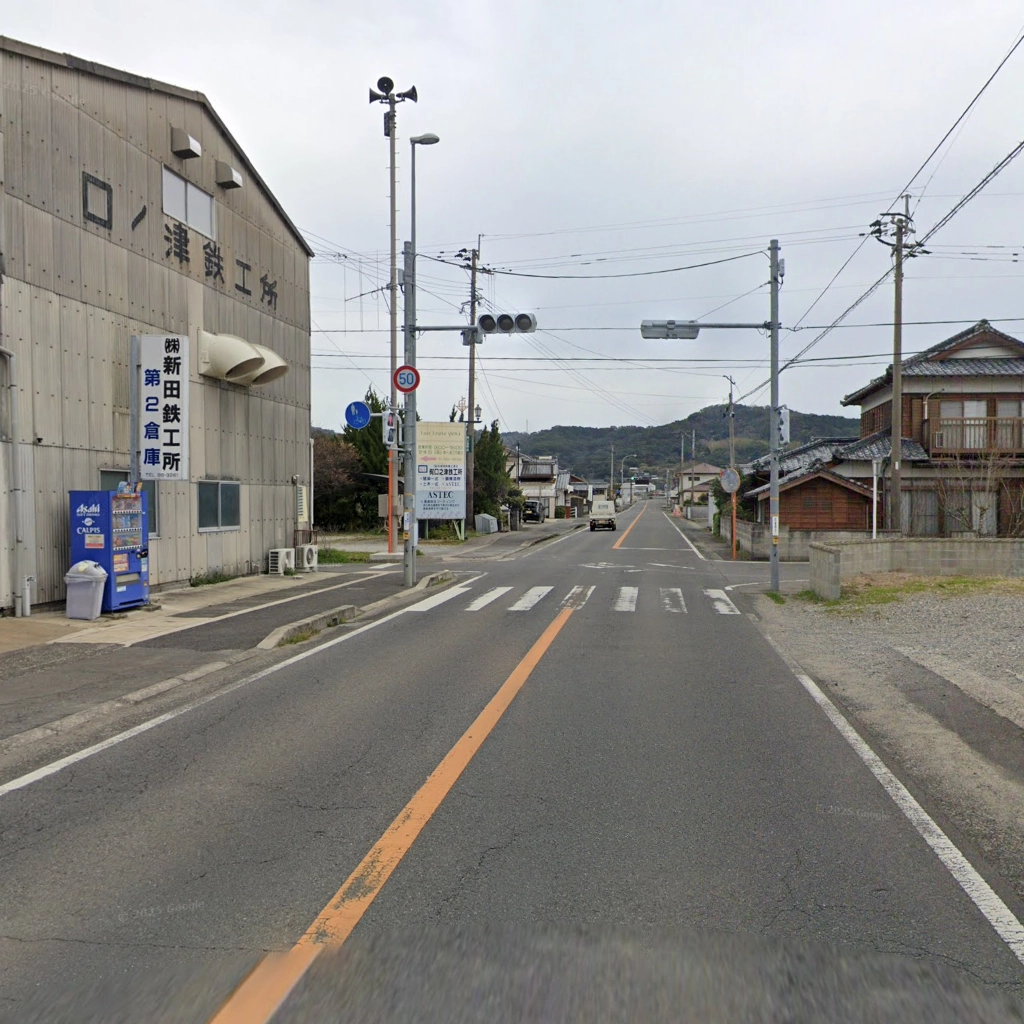 曇り空の下、信号機のある交差点の風景。左手前には「(株)新田鉄工所 第2倉庫」と書かれた看板と青い自動販売機がある大きな工場が建っている。道路は中央線がオレンジ色。この信号で加津佐方面からは右折、南有馬方面からは左折する。