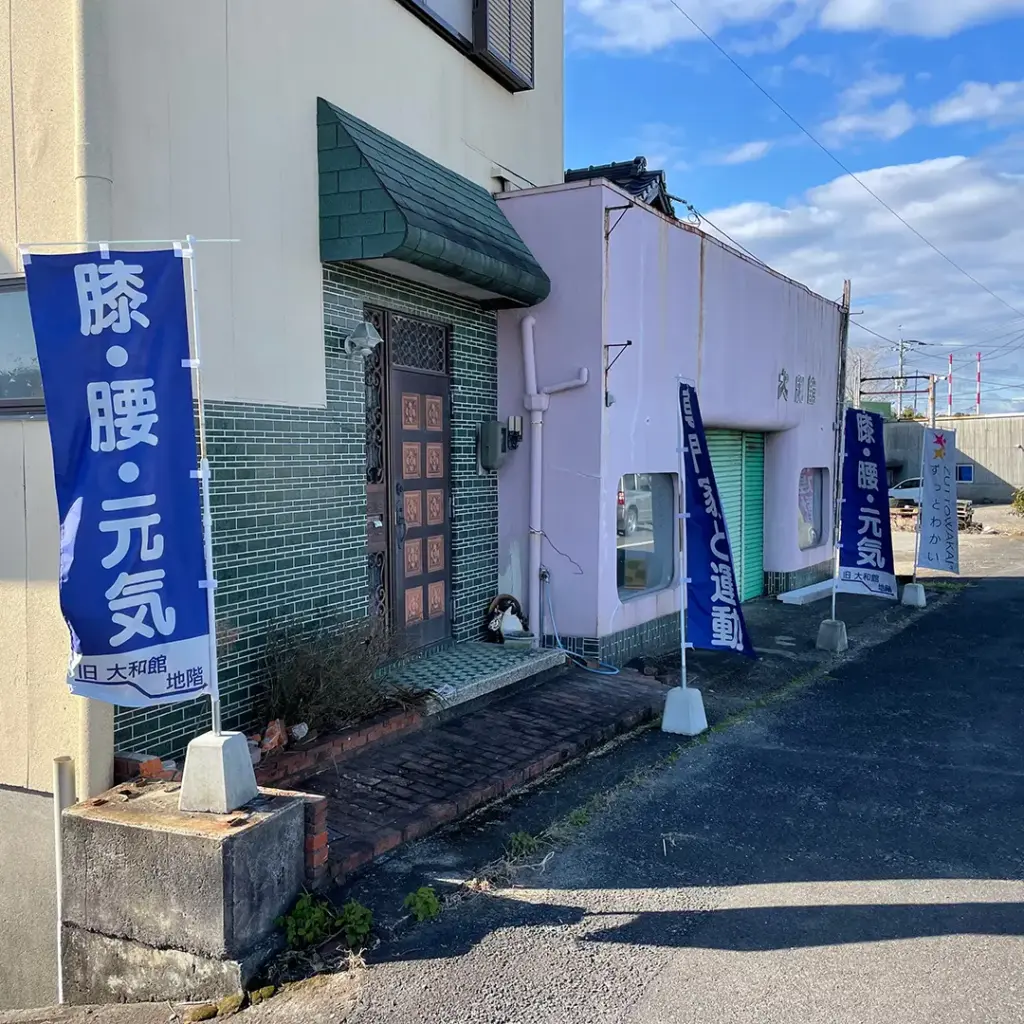 佐賀県太良町のジム 健康運動ZUTTOWAKAIの外観。国道沿いの「旧大和館」の地階にあり、「膝・腰・元気」と書かれた青いノボリが目印です。地域の皆様が気軽に立ち寄り、安心して通えるアットホームな入り口の様子を紹介しています。