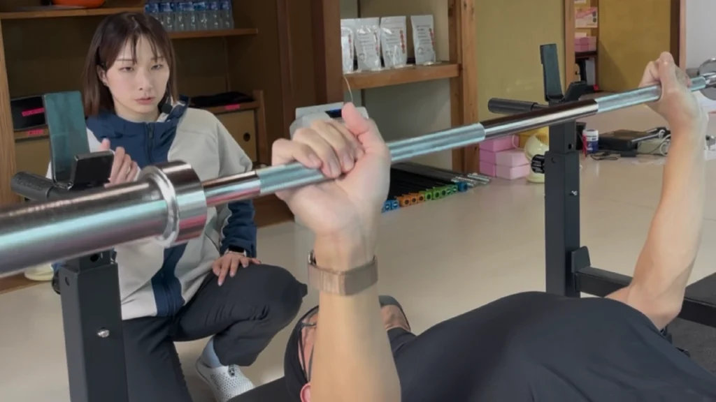 ZUTTOWAKAIのジムで、女性トレーナーがベンチプレスを行う会員のフォームを確認しながら指導している職場のトレーニング風景